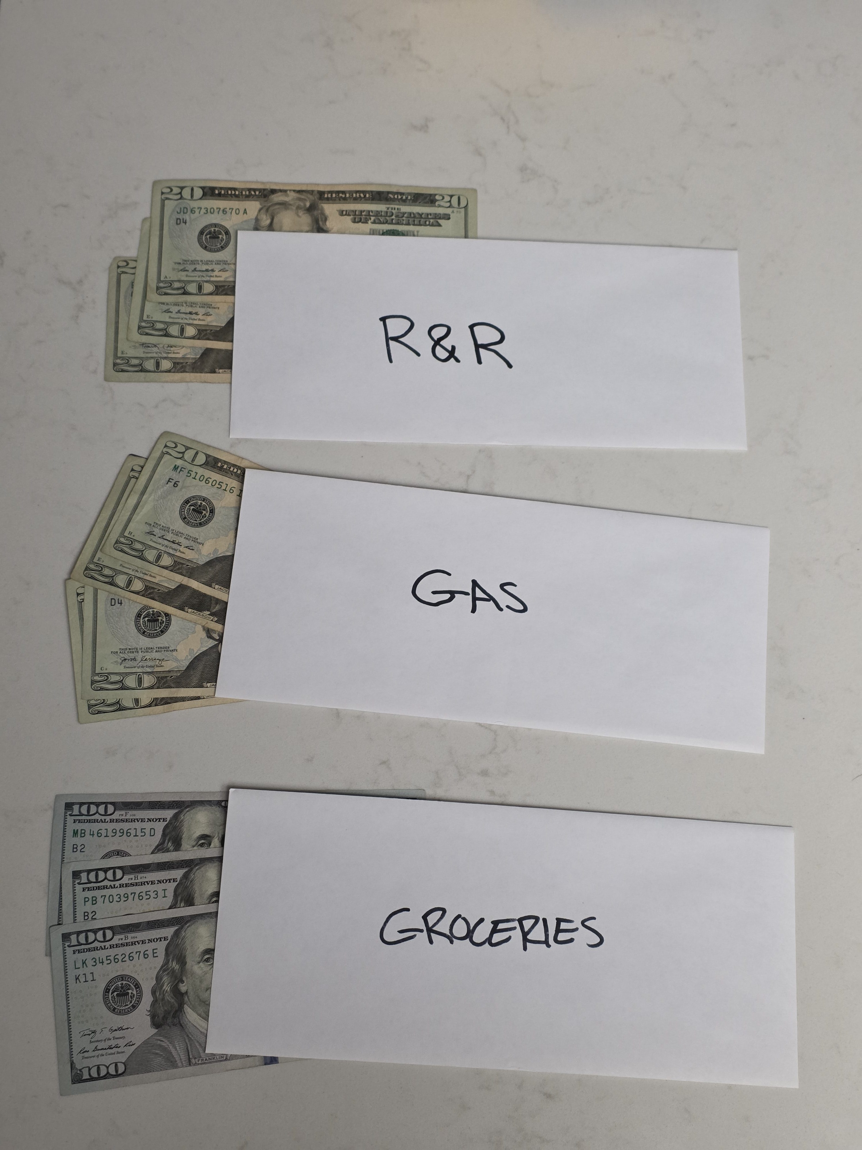 Three white envelopes labeled R&R, Gas, and Groceries with cash peeking out, representing a physical envelope budgeting system on a kitchen counter.